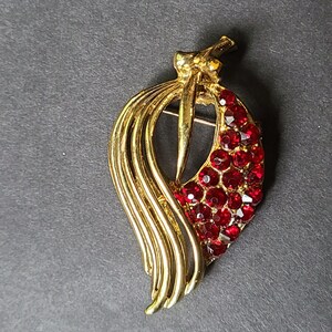 May include: A vintage gold-tone brooch in a leaf design, adorned with sparkling red rhinestones. The brooch features a central pin and a flowing, layered design, creating a classic and elegant accessory.