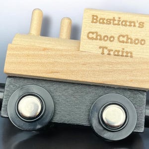 Op de afbeelding: Houten speelgoedtrein met de tekst "Bastian's Choo Choo Train" gegraveerd op de bovenkant. De trein heeft een bovenkant in natuurlijke houtkleur en een zwarte basis met twee zwarte wielen. De trein heeft twee kleine houten schoorstenen.