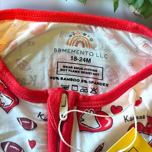 K&ouml;nnte beinhalten: Wei&szlig;-roter Baby-Schlafanzug mit Rei&szlig;verschluss. Das Muster zeigt Footballs und Herzen, mit dem Schriftzug "Kansas City". Der Halsausschnitt und die Verzierungen sind rot. Das Etikett lautet "BBMEMENTO LLC 18-24M".