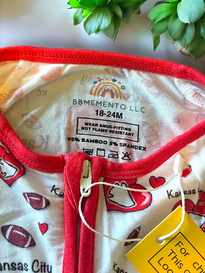 K&ouml;nnte beinhalten: Wei&szlig;-roter Baby-Schlafanzug mit Rei&szlig;verschluss. Das Muster zeigt Footballs und Herzen, mit dem Schriftzug "Kansas City". Der Halsausschnitt und die Verzierungen sind rot. Das Etikett lautet "BBMEMENTO LLC 18-24M".