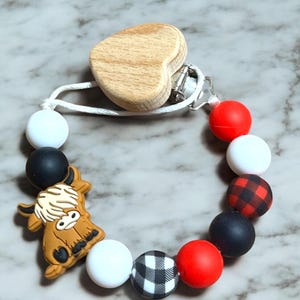 May include: A silicone baby teether with a wooden heart clip. The teether features a red, white, and black buffalo plaid pattern, a white bead, a black bead, a red bead, and a brown and white highland cow charm.