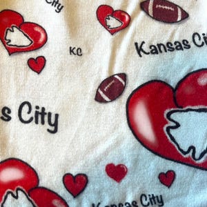 K&ouml;nnte beinhalten: Wei&szlig;er Stoff mit einem sich wiederholenden Muster aus roten Herzen, Footballs und den Worten "Kansas City" in Schwarz. Einige Herzen enthalten ein wei&szlig;es Pfeilspitzendesign. Die Buchstaben "kc" sind ebenfalls vorhanden.