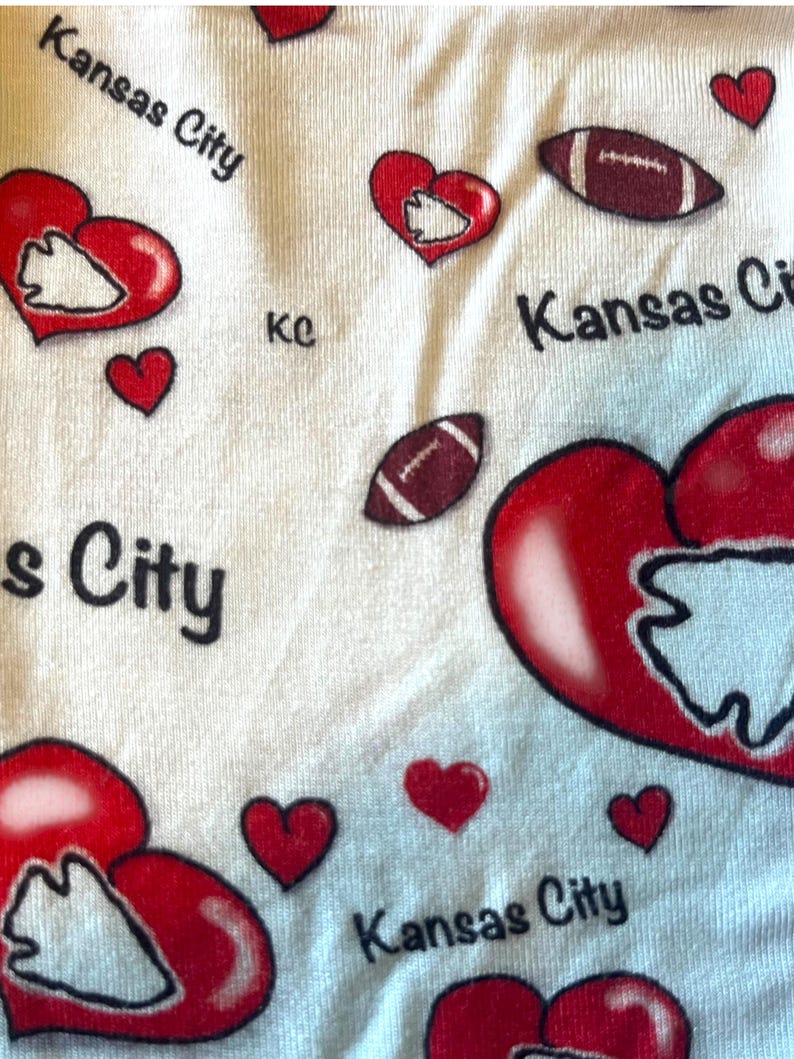 K&ouml;nnte beinhalten: Wei&szlig;er Stoff mit einem sich wiederholenden Muster aus roten Herzen, Footballs und den Worten "Kansas City" in Schwarz. Einige Herzen enthalten ein wei&szlig;es Pfeilspitzendesign. Die Buchstaben "kc" sind ebenfalls vorhanden.