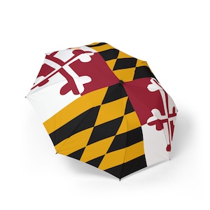 May include: An open umbrella featuring the Maryland state flag design. The umbrella has a white, red, yellow, and black color scheme with a repeating pattern of crosses and diamond shapes. The umbrella is designed to provide protection from the rain.