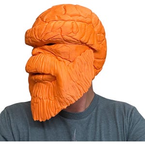 May include: Orange mask with a textured, rocky appearance, covering the head and face. The mask features a prominent brow, nose, and a detailed beard. The person is wearing a gray t-shirt.