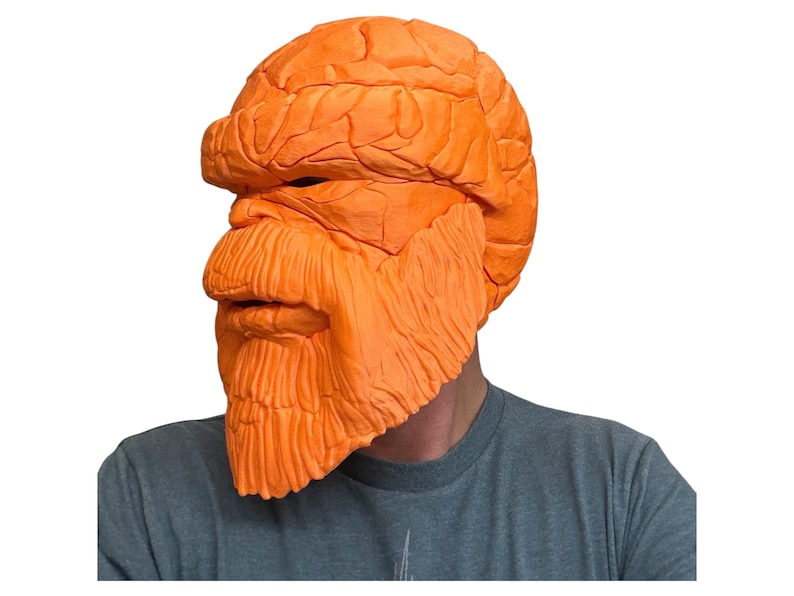 May include: Orange mask with a textured, rocky appearance, covering the head and face. The mask features a prominent brow, nose, and a detailed beard. The person is wearing a gray t-shirt.