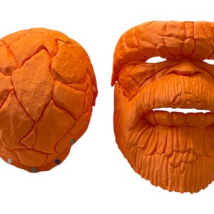 May include: Two orange masks, one spherical and the other a detailed face. The face mask has a textured surface, with eye and mouth openings, and a prominent beard. The spherical mask has a segmented, rock-like appearance. Both masks are brightly colored.