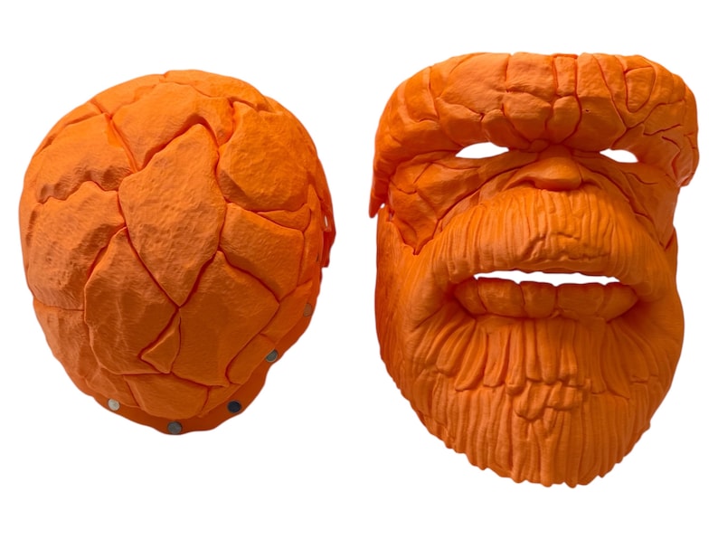 May include: Two orange masks, one spherical and the other a detailed face. The face mask has a textured surface, with eye and mouth openings, and a prominent beard. The spherical mask has a segmented, rock-like appearance. Both masks are brightly colored.