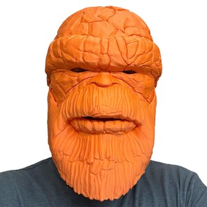 May include: Orange mask resembling a character with a textured, rocky appearance. The mask features a detailed face with a prominent brow, nose, and beard. The mask is worn by a person wearing a gray shirt. A costume or cosplay item.