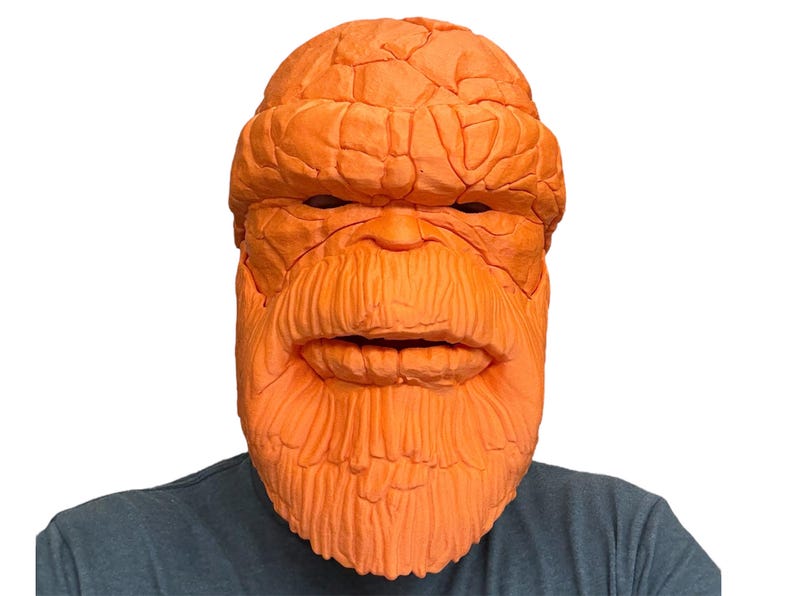 May include: Orange mask resembling a character with a textured, rocky appearance. The mask features a detailed face with a prominent brow, nose, and beard. The mask is worn by a person wearing a gray shirt. A costume or cosplay item.