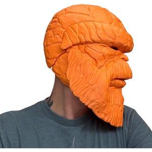 May include: An orange mask with a textured, rocky appearance. The mask covers the head and face, featuring a prominent brow, nose, and beard-like structure. The mask is a solid orange color and appears to be made of a rigid material.