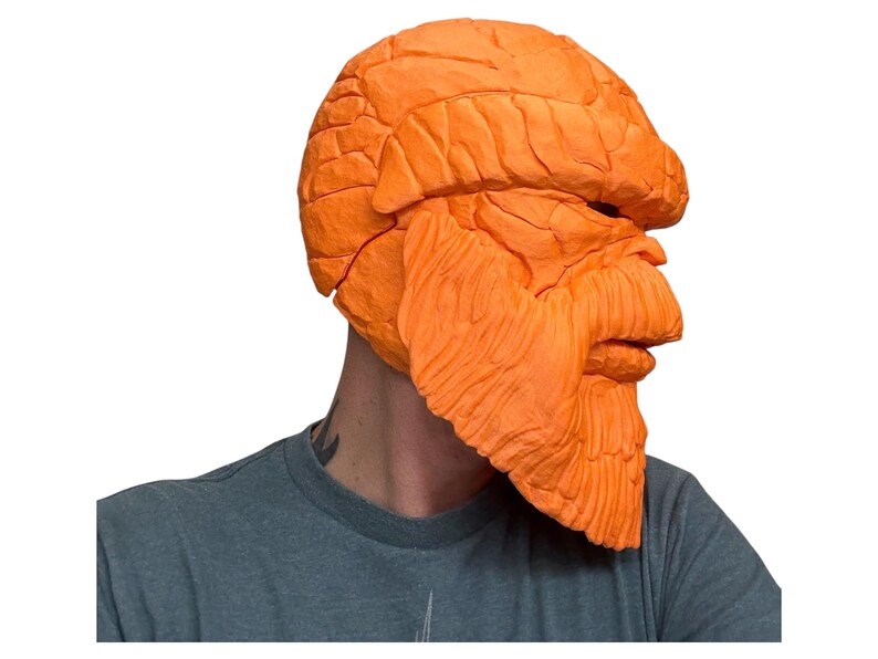 May include: An orange mask with a textured, rocky appearance. The mask covers the head and face, featuring a prominent brow, nose, and beard-like structure. The mask is a solid orange color and appears to be made of a rigid material.