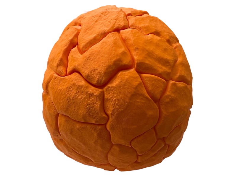 May include: An orange, spherical object with a textured, segmented surface. The segments are irregular and give the object a rough, organic appearance. The object is set against a plain white background.