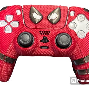 May include: A red and black PlayStation 5 controller with a Spiderman design. The controller has a textured surface and a black and white Spiderman eye design on the top.