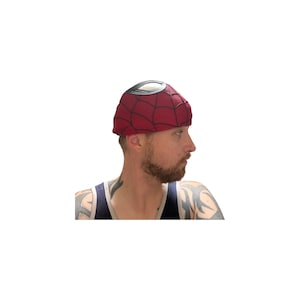May include: A red beanie-style hat with a black web pattern and a white and black eye design. The hat is a costume accessory, likely inspired by a superhero. The hat is made of a soft material.