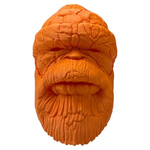 May include: Orange sculpted mask resembling a character with a textured, rocky appearance. The mask features a detailed face with a furrowed brow, a wide mouth, and a prominent chin. The mask is likely for cosplay or costume use.
