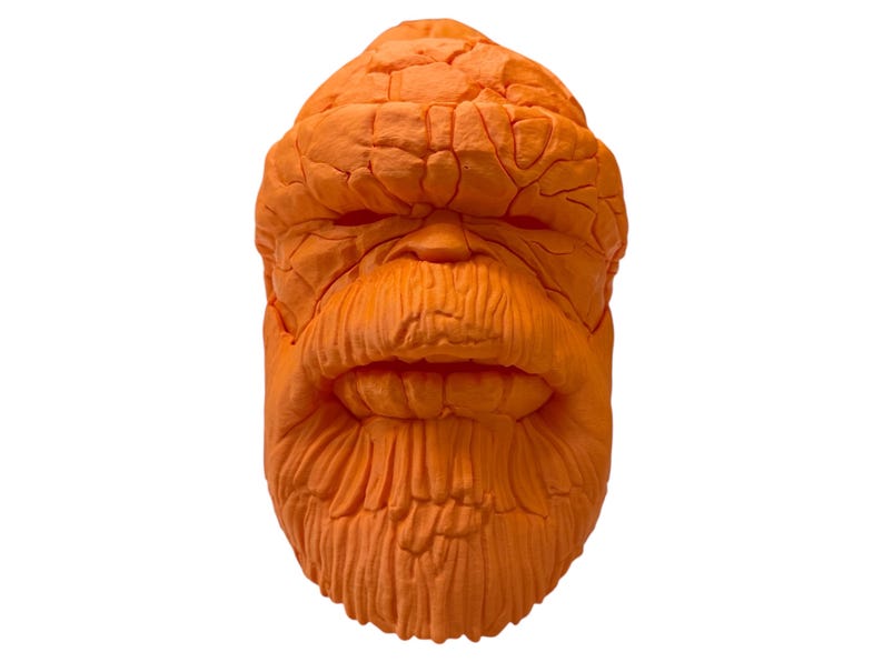 May include: Orange sculpted mask resembling a character with a textured, rocky appearance. The mask features a detailed face with a furrowed brow, a wide mouth, and a prominent chin. The mask is likely for cosplay or costume use.