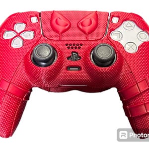 May include: A red PlayStation 5 DualSense controller with a textured surface and a Spider-Man design. The controller has a black PlayStation logo and black buttons.