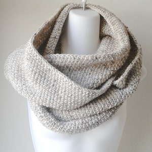May include: A crocheted infinity scarf in a light beige color. The scarf is made with a textured stitch and is displayed on a white mannequin.