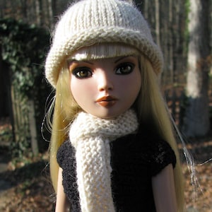 May include: A doll with blonde hair wearing a white knitted hat and scarf, and a black dress. The doll has brown eyes and a neutral expression.