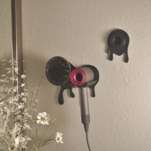 May include: Black wall-mounted hair dryer holder with a unique drip design. The holder is designed to accommodate a hair dryer with a pink and silver finish. A matching holder for the diffuser is also mounted on the wall.