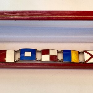 May include: A silver bracelet with enamel nautical flags. The flags are red, white, blue, and yellow. The bracelet is in a red velvet box.
