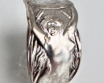 May include: A silver-toned metal ring featuring a raised relief design of a figure with long flowing hair, arms raised above the head, and textured details along the sides. The band has a polished finish with dark recessed areas.