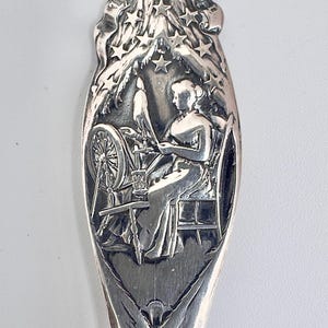 May include: A silver-toned metal spoon handle featuring an embossed scene of a person sitting at a spinning wheel. The design includes a starry sky motif above the figure, with intricate decorative carvings extending down the length of the handle.
