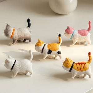Può includere: Cinque piccole statuette di gatti in vari colori e motivi, ognuna con un semplice collare. La collezione comprende un gatto grigio con collare arancione, un gatto bianco con collare nero, un gatto calico, un gatto rosa e bianco e un gatto tigrato.