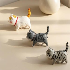 Può includere: Tre piccole statuette di gatti in fila. Una è bianca con macchie arancioni e gialle e un collare rosso. La seconda è grigio scuro con strisce nere e un collare scuro. La terza è grigio chiaro con strisce nere e un collare scuro.