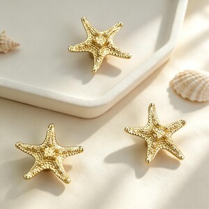 May include: Three gold-toned metal drawer knobs shaped like five-pointed starfish. Each knob features a textured, bumpy surface detail mimicking the natural appearance of a starfish. The knobs are arranged with two positioned below and one above.