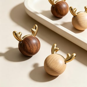 May include: A collection of spherical cabinet knobs featuring a smooth wooden base in dark walnut or light oak finishes, topped with a polished gold-toned metal accent shaped like a pair of deer antlers, providing a functional grip for furniture.
