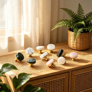 May include: A collection of decorative cabinet knobs in various colors and patterns. The knobs feature marble, onyx, and other stone materials with gold-toned bases. The knobs are displayed on a light brown woven surface.