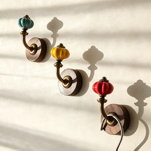 May include: Three wall-mounted hooks featuring round dark wood bases, curved brass-toned metal arms, and fluted ceramic knobs in light blue, yellow, and red. The bottom hook includes a silver-toned metal S-hook hanging from the brass arm.