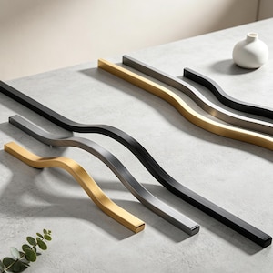 May include: A collection of six long, wavy cabinet handles arranged in parallel. The set includes two matte black, two brushed gold, and two brushed silver metal pulls, each featuring a distinct curved design and a rectangular profile.