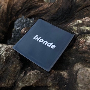 May include: A dark blue rectangular object with the word "blonde" in white lettering. The object is reflective and sits on a textured, brown wooden surface. The font is a clean, sans-serif style. The overall aesthetic is minimalist.