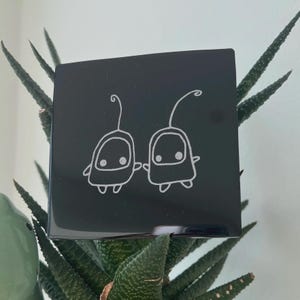 May include: A black square with a white line drawing of two cartoon characters. The characters have oval bodies, small eyes, and antennae. The square is resting on a green plant with spiky leaves.