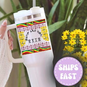 May include: White travel mug with a handle, featuring a decorative band with Ethiopian flag colors and text. The mug is held in front of a background of yellow flowers and greenery.