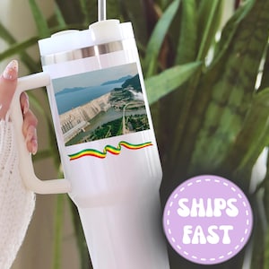 May include: White travel mug with a handle, featuring a landscape image of a dam and a colorful ribbon design. The mug has a white lid and a straw. The mug is held by a person.