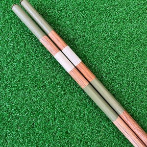 Könnte beinhalten: Zwei Holzstäbchen mit einem grün-weiß-braunen Streifenmuster. Die Stäbchen liegen auf einer grünen Kunstgrasoberfläche.