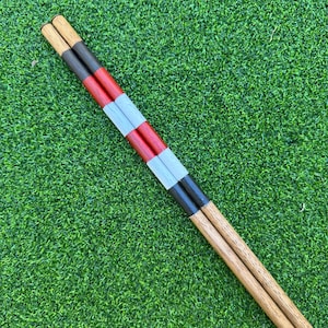 Peut inclure: Deux cravaches de dressage en bois avec des bandes alternées de peinture noire, rouge et argentée. Les cravaches ont une finition en bois naturel sur les poignées et sont posées sur un fond vert.