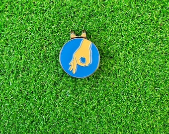 May include: A blue and yellow golf ball holder with a hand making the "OK" gesture.