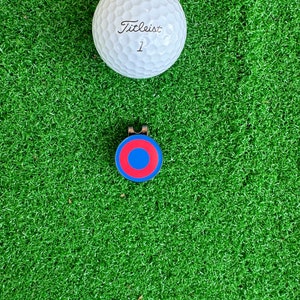 May include: A white golf ball with the text "Titleist 1" printed on it, and a blue and red golf ball marker.