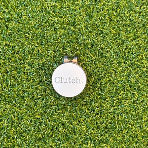 May include: A white golf ball marker with a silver clip and the word "Clutch." printed on it. The marker is on a green artificial grass surface.