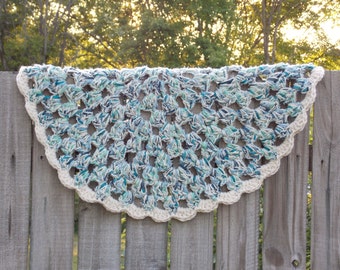May include: A crocheted shawl in shades of blue and cream, draped over a wooden fence. The shawl has an openwork design with a scalloped cream border. The yarn has a textured appearance.