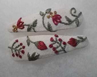 May include: Two white fabric hair clips with embroidered floral designs. The embroidery features red flowers, green leaves, and grey stems. The clips are oval-shaped and have a textured appearance.