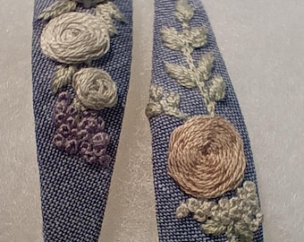 May include: A pair of denim-effect hair clips, each with embroidered floral patterns. One clip has three light grey roses and a cluster of purple grapes. The other features a single brown rose and green leaves.