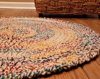 May include: A circular, hand-knitted rug with a spiral pattern. The rug incorporates a variety of colours, such as cream, yellow, teal, red, and brown. The rug is positioned on a wooden floor, with a woven basket visible in the background.
