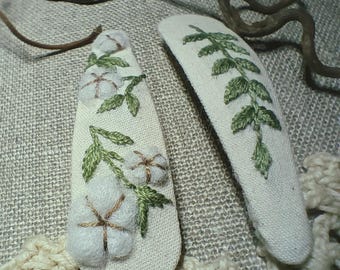 May include: Two off-white fabric hair clips. One clip features embroidered cotton flowers and green leaves. The other clip has a green leaf design. The clips are on a textured, neutral-toned surface.