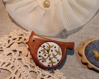 May include: A wooden bird brooch with an embroidered circular design of green leaves and blue berries. The brooch is set against a backdrop of lace and linen fabric, showcasing a handcrafted aesthetic.